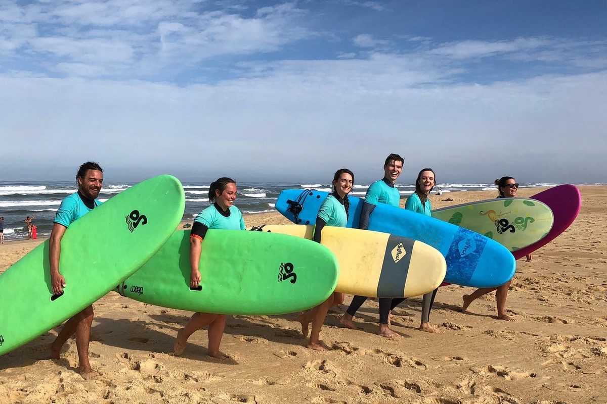 Des élèves de surf souriants portent leur planche le long de la plage.